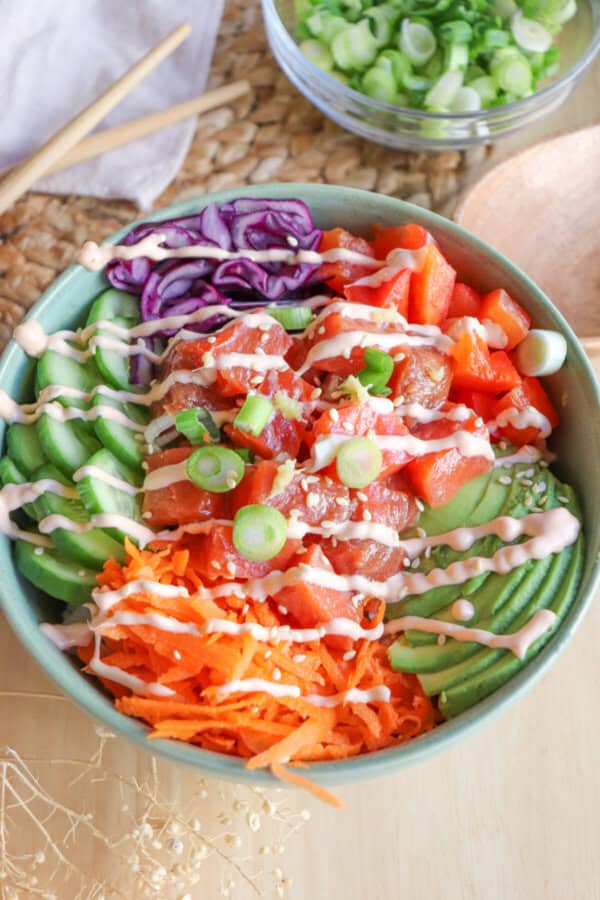 A green bowl with sliced cucumbers, grated carrot, sliced avocado, purple cabbage, diced red pepper with raw cubes of salmon topped with a drizzle of spicy mayo and diced green onion.