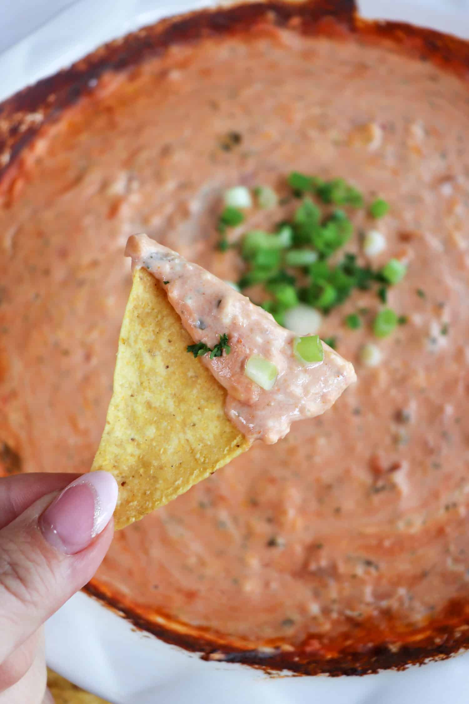 A tortilla chip with dip held in a hand over a dish of cheesy salsa dip topped with diced green onions.