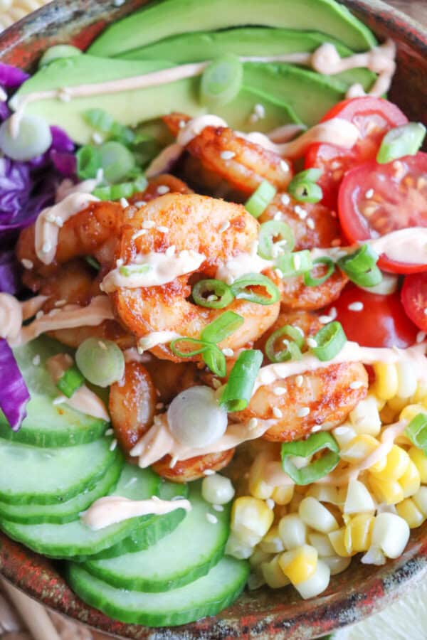 Cooked shrimp in a bowl surrounded by sliced cucumber, corn kernels, cherry tomatoes, sliced avocado and shredded red cabbage drizzled with spicy mayonnaise.