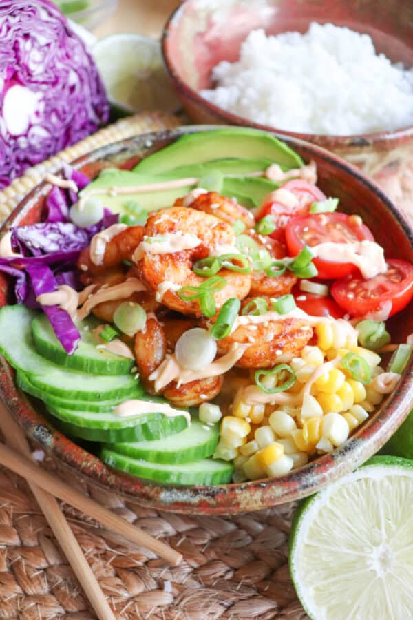 Cooked shrimp in a bowl surrounded by sliced cucumber, corn kernels, cherry tomatoes, sliced avocado and shredded red cabbage drizzled with spicy mayonnaise.