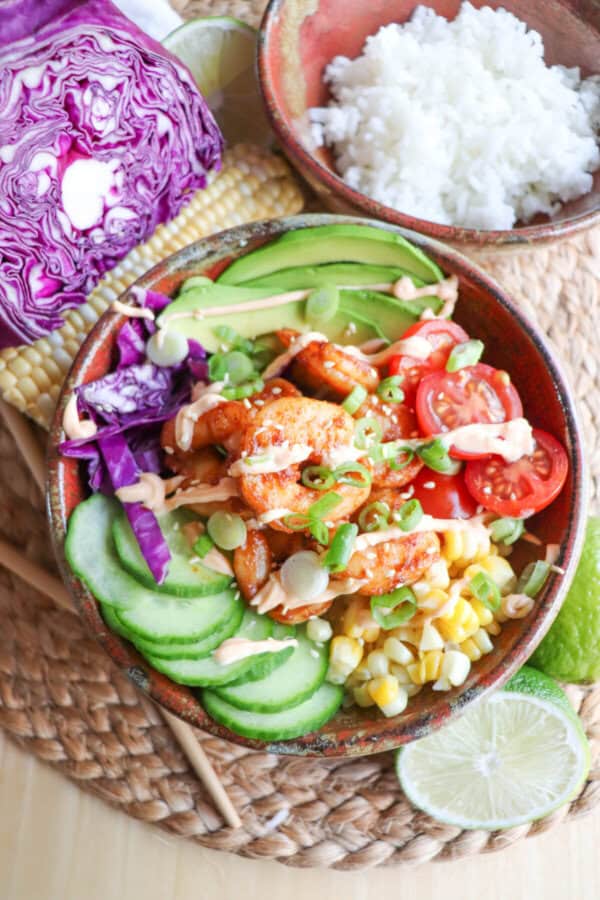 Cooked shrimp in a bowl surrounded by sliced cucumber, corn kernels, cherry tomatoes, sliced avocado and shredded red cabbage drizzled with spicy mayonnaise.