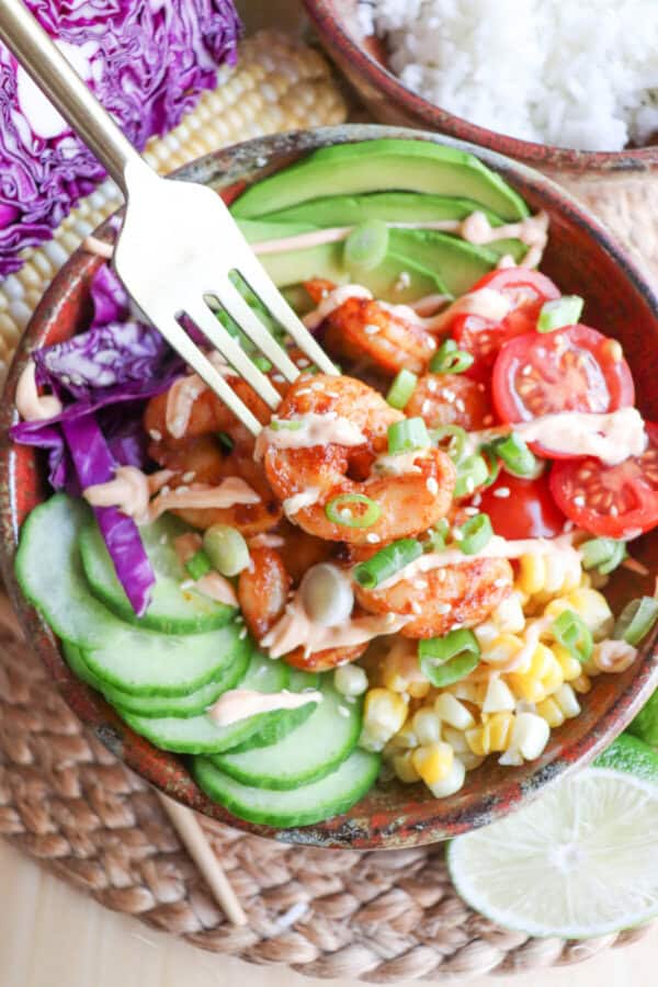 Cooked shrimp in a bowl surrounded by sliced cucumber, corn kernels, cherry tomatoes, sliced avocado and shredded red cabbage drizzled with spicy mayonnaise and a shrimp on a fork.