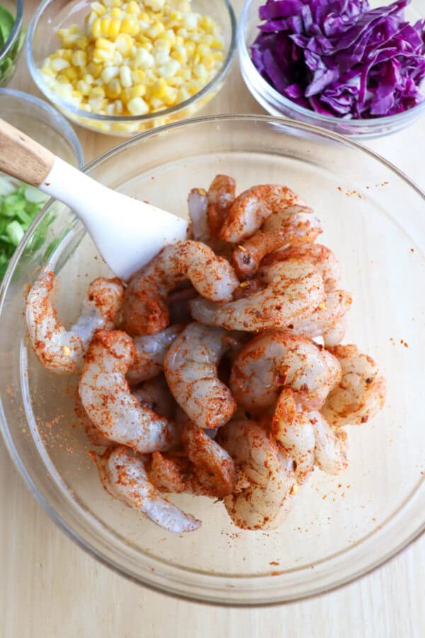 Raw shrimp in a clear glass bowl coated in seasonings with a white spatula in the bowl.