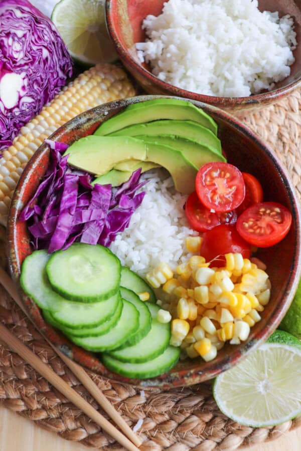 A brown bowl filled with rice with a selection of vegetables including corn kernels, cherry tomatoes, sliced avocado, red cabbage and sliced cucumbers around the edge.