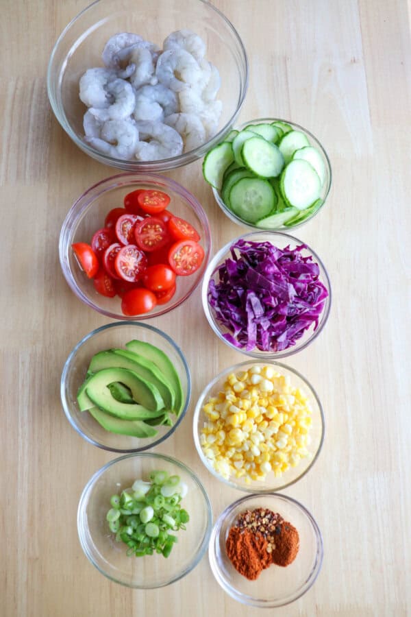 Ingredients for shrimp poke in clear glass bowls on a wood table.