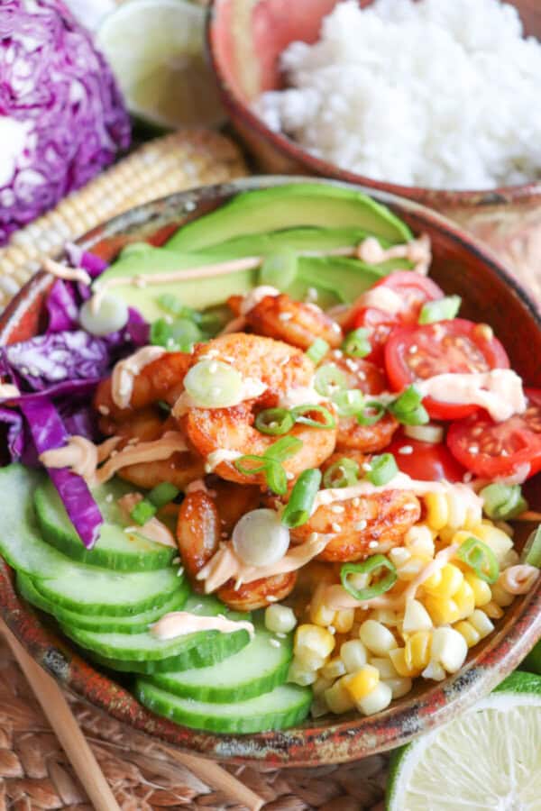 Cooked shrimp in a bowl surrounded by sliced cucumber, corn kernels, cherry tomatoes, sliced avocado and shredded red cabbage drizzled with spicy mayonnaise.