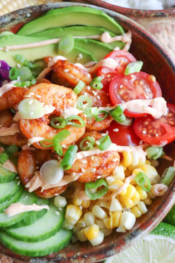 Close up of cooked shrimp in a bowl surrounded by sliced cucumber, corn kernels, cherry tomatoes, sliced avocado and shredded red cabbage drizzled with spicy mayonnaise.