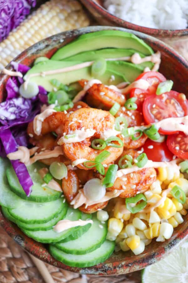 Cooked shrimp in a bowl surrounded by sliced cucumber, corn kernels, cherry tomatoes, sliced avocado and shredded red cabbage drizzled with spicy mayonnaise.