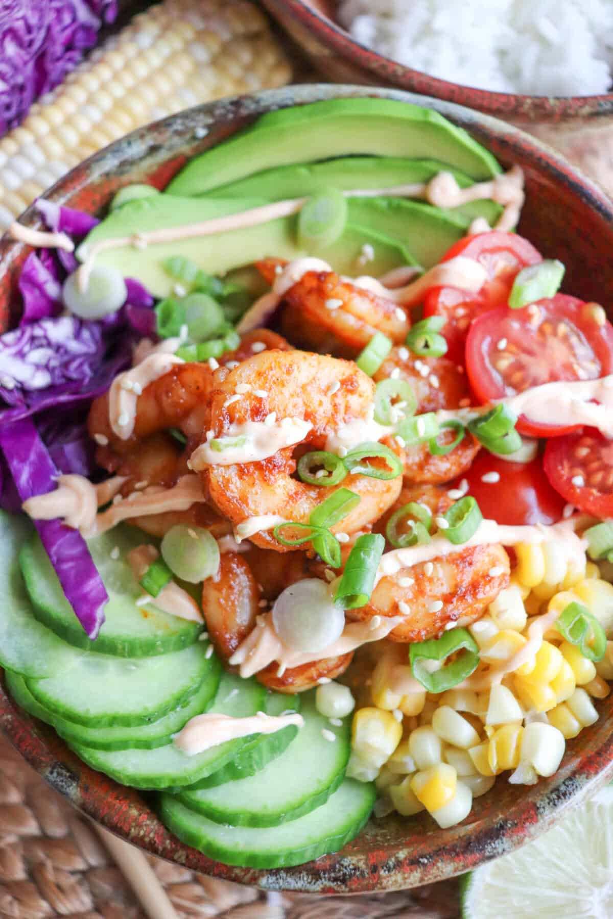 Cooked shrimp in a bowl surrounded by sliced cucumber, corn kernels, cherry tomatoes, sliced avocado and shredded red cabbage drizzled with spicy mayonnaise.