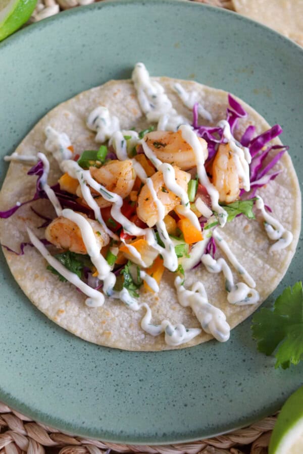 White corn tortilla on a green plate layered with red cabbage, fresh salsa, cooked shrimp and a drizzle of crema.