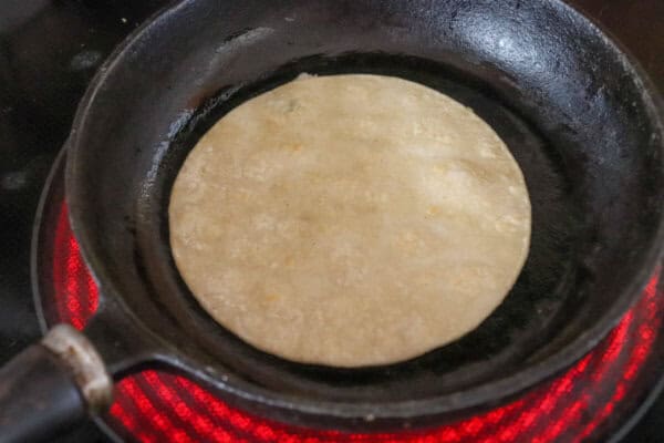 Four inch white corn tortilla in a cast iron skillet on the stove top.