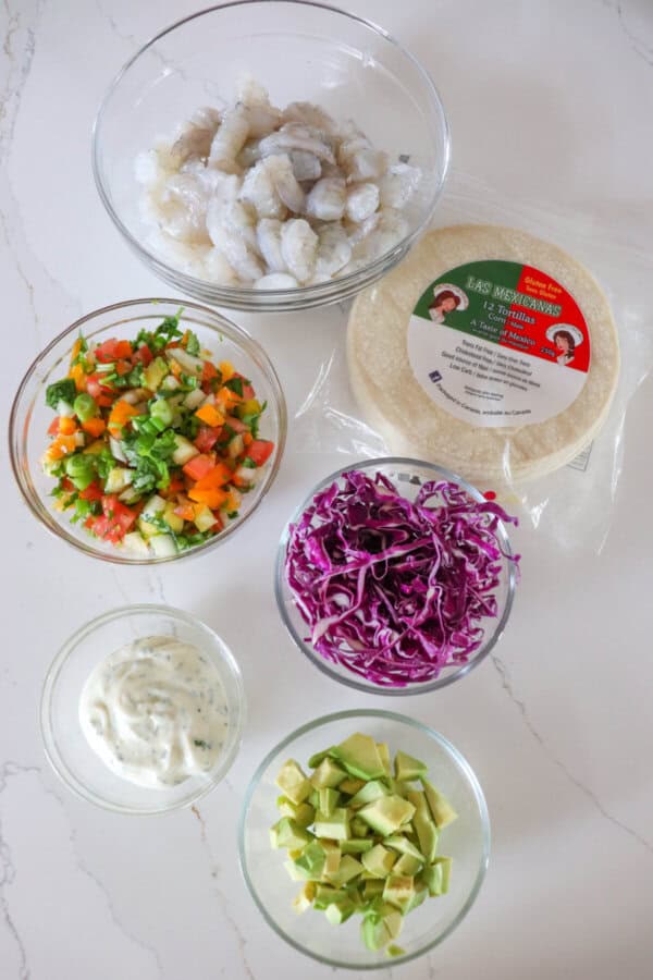 Shrimp taco ingredients in clear glass bowls on a white counter.