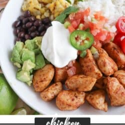 Cooked chicken bites with seasoning in a large white bowl with rice, diced avocado, black beans, corn kernels, cherry tomatoes, pico and sour cream topped with a slice of jalapeno and lime.