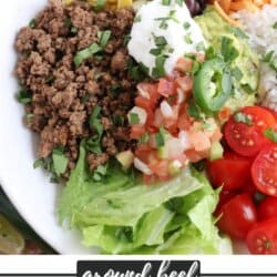 Large white bowl filled with cooked ground taco beef, rice, pico, corn, beans, cheddar cheese and cherry totmatoes, topped with guacamole and sour cream.