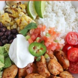 Cooked chicken bites with seasoning in a large white bowl with rice, diced avocado, black beans, corn kernels, cherry tomatoes, pico and sour cream topped with a slice of jalapeno and lime.