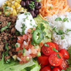 Large white bowl filled with cooked ground taco beef, rice, pico, corn, beans, cheddar cheese and cherry totmatoes, topped with guacamole and sour cream.