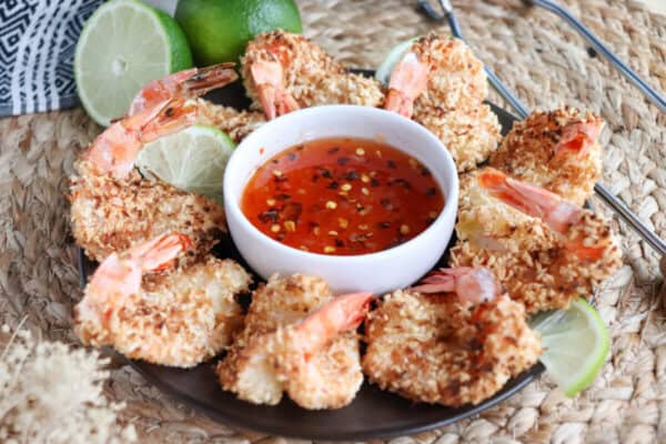 Sweet chili red sauce with red pepper chili flakes in a small white bowl surrounded by coconut shrimp.