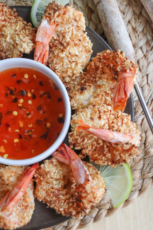 Close up of sweet chili red sauce with red pepper chili flakes in a small white bowl surrounded by coconut shrimp.