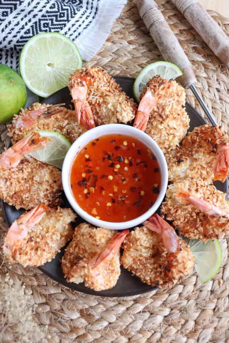 Sweet chili red sauce with red pepper chili flakes in a small white bowl surrounded by coconut shrimp.