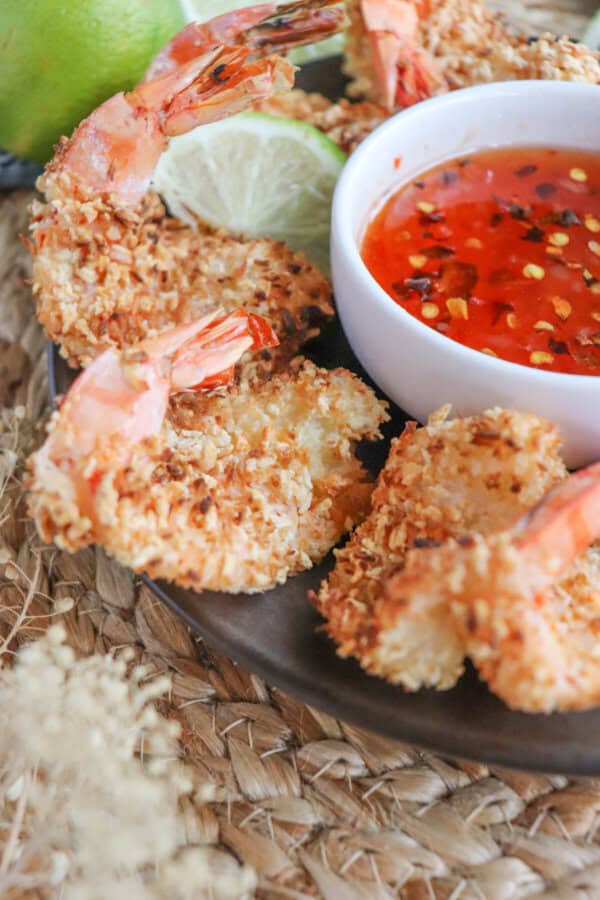 Close up of sweet chili red sauce with red pepper chili flakes in a small white bowl surrounded by coconut shrimp.