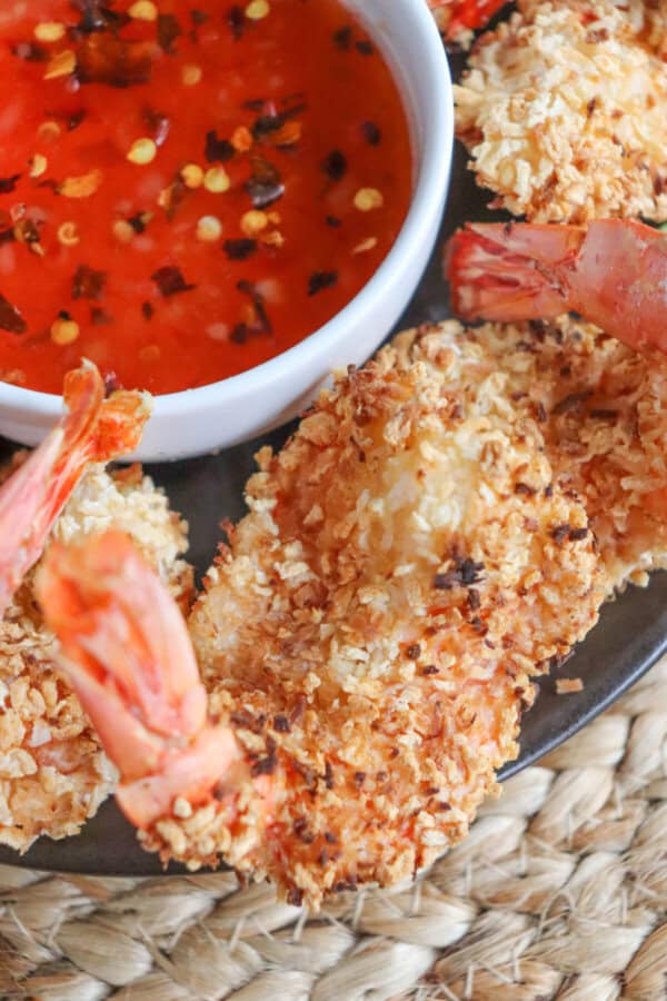 Close up of sweet chili red sauce with red pepper chili flakes in a small white bowl surrounded by coconut shrimp.