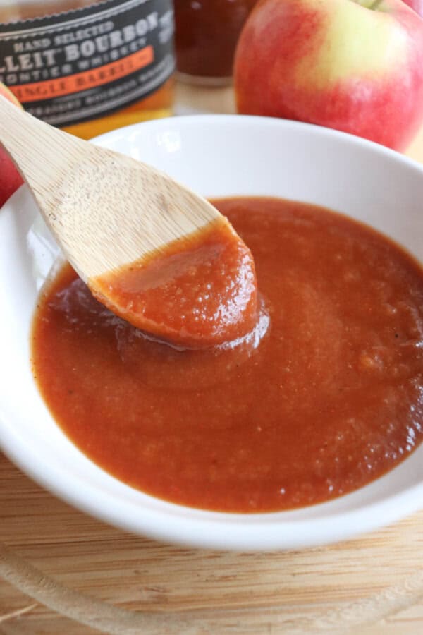 Apple bourbon BBQ sauce in a small white bowl with a wooden spoon dipping into it.
