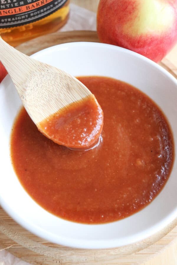 Apple bourbon BBQ sauce in a small white bowl with a wooden spoon dipping into it.