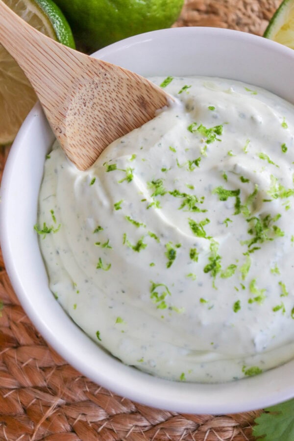 White bowl of crema sauce topped with lime zest with a wooden spoon dipped in on the left.