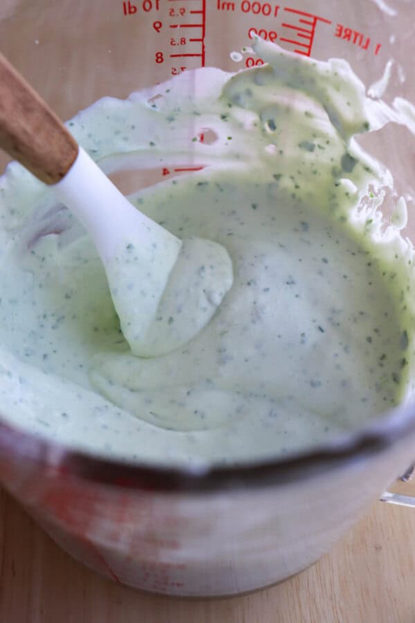 Creamy white crema sauce with green tint and green cilantro bits in a pyrex measuring cup with a white spatula in it.