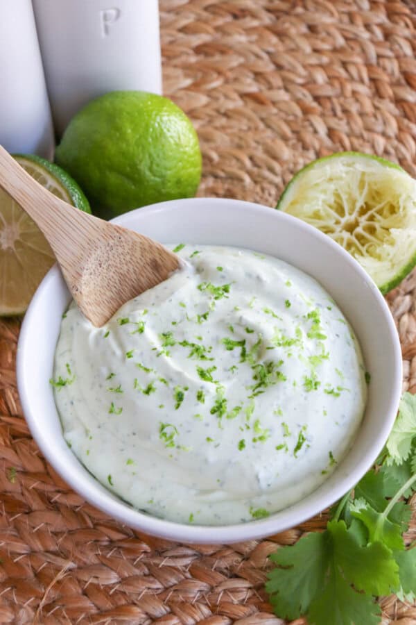White bowl of crema sauce topped with lime zest with a wooden spoon dipped in on the left.