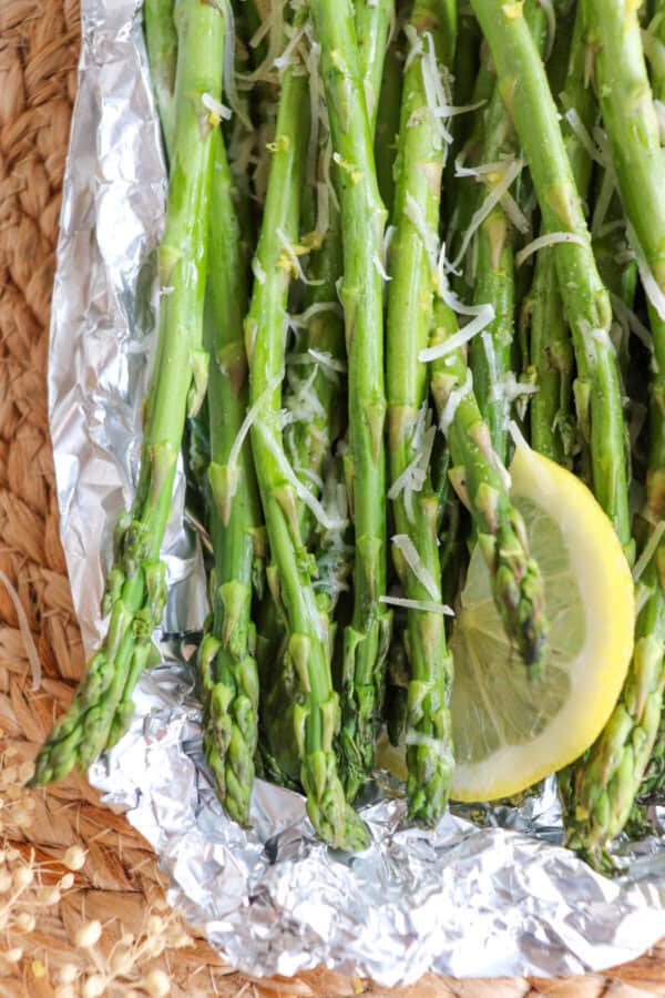 Close up of cooked asparagus in a foil packed topped with shredded parmesan cheese and lemon slices.