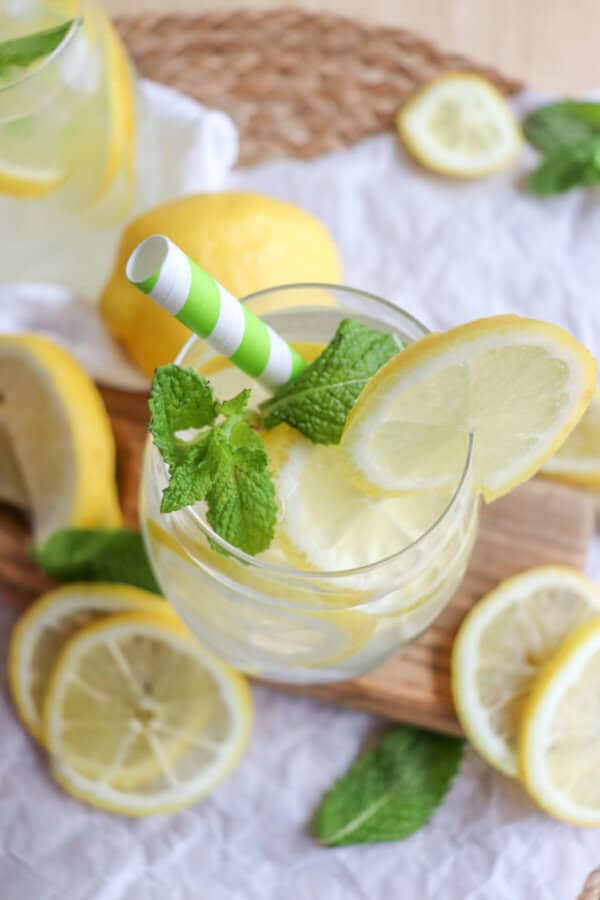 Limoncello spritz in a glass with lemon slices and mint leaves with a green and white straw surrounded by lemon slices on a wooden cutting board.