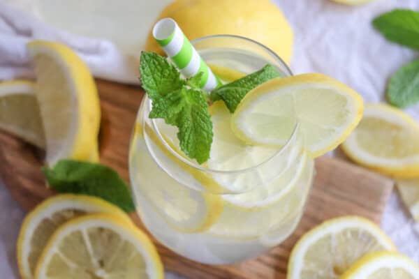Limoncello spritz in a glass with lemon slices and mint leaves with a green and white straw surrounded by lemon slices on a wooden cutting board.