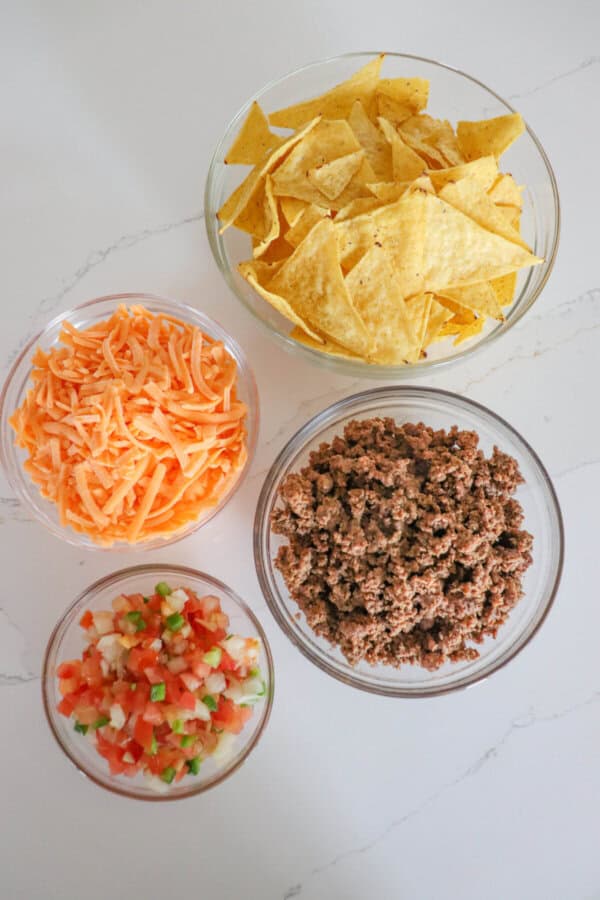 Ingredients for air fryer nachos in clear glass bowls on a white counter.