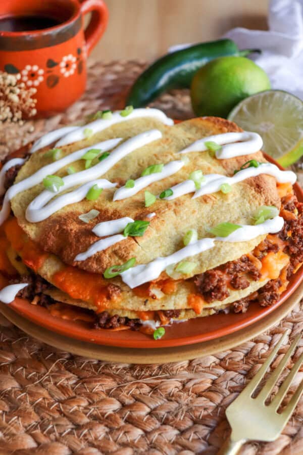 Corn tortillas layered with meat and cheese and topped with lime crema and sliced green onion on an orange plate.