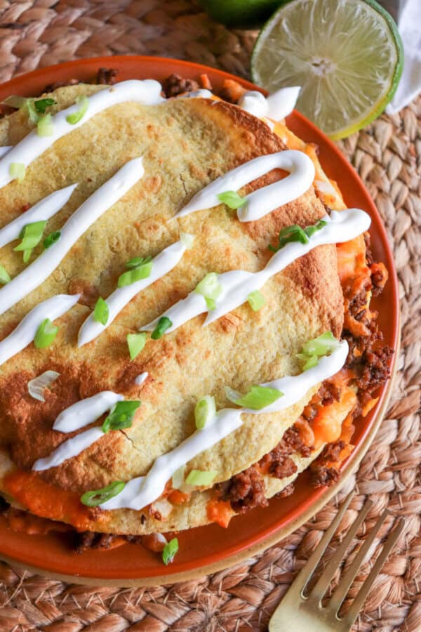 Four inch corn tortillas layered with meat and cheese and topped with lime crema and sliced green onion on an orange plate.