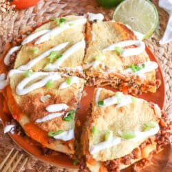 Four inch corn tortillas layered with meat and cheese and topped with lime crema and sliced green onion on an orange plate.