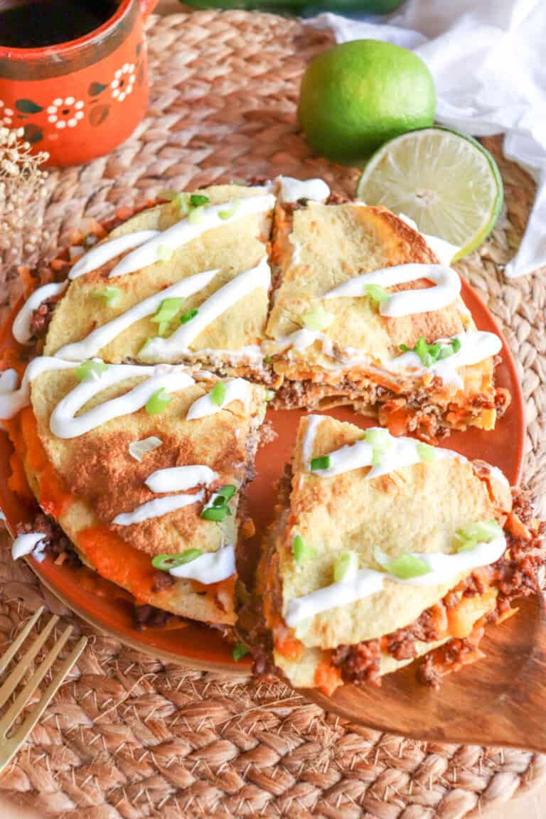 Four inch corn tortillas layered with meat and cheese and topped with lime crema and sliced green onion on an orange plate.