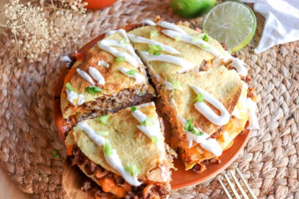 Four inch corn tortillas layered with meat and cheese and topped with lime crema and sliced green onion on an orange plate.