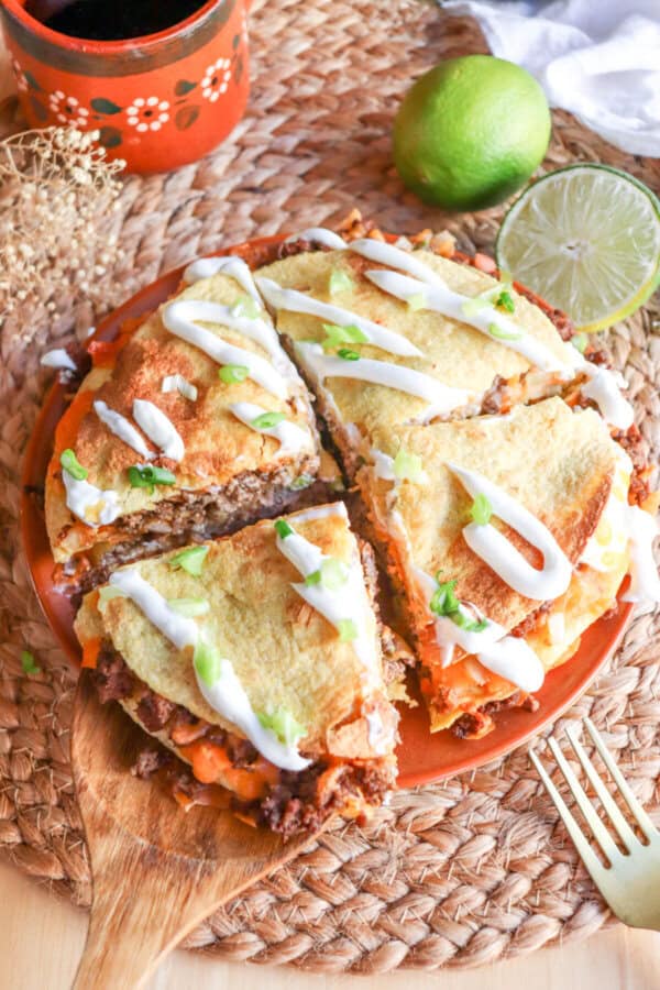 Corn tortillas layered with meat and cheese and topped with lime crema and sliced green onion on an orange plate.