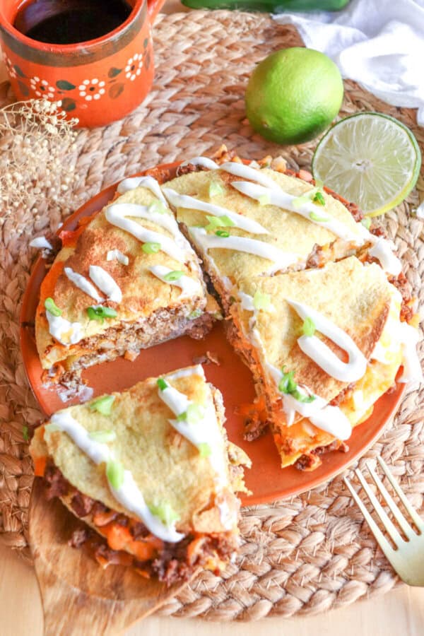 Corn tortillas layered with meat and cheese and topped with lime crema and sliced green onion on an orange plate.