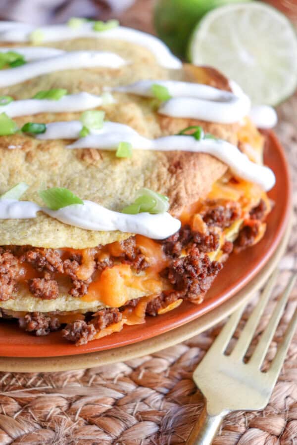 Close up of corn tortillas layered with meat and cheese and topped with lime crema and sliced green onion on an orange plate.
