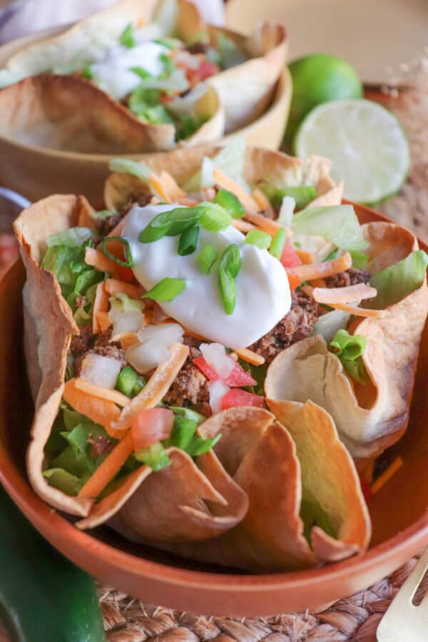 Taco salad topped with sour cream & green onions in a tortilla bowl in an orange bowl on a wooden table.
