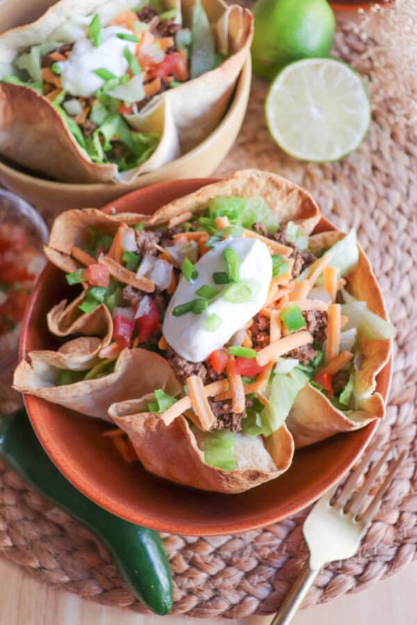 Taco salad topped with sour cream & green onions in a tortilla bowl in an orange bowl on a wooden table.
