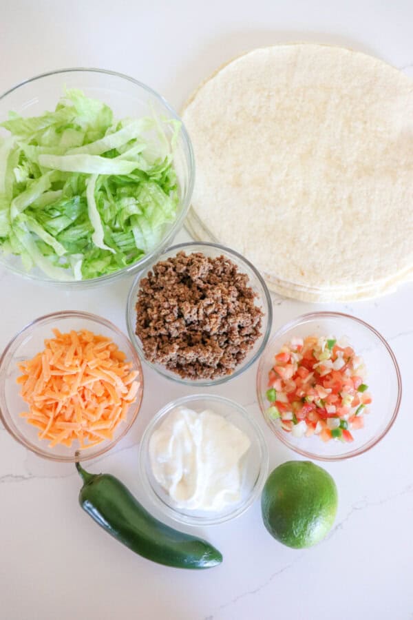 Ingredients for air fryer taco salad in clear glass bowls on a white counter.
