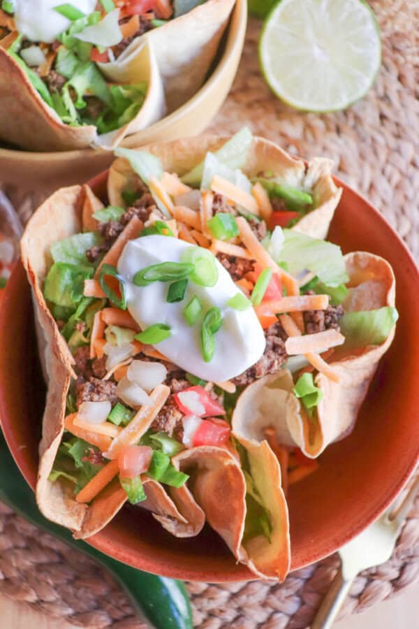 Taco salad topped with sour cream & green onions in a tortilla bowl in an orange bowl on a wooden table.