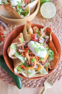 Taco salad topped with sour cream & green onions in a tortilla bowl in an orange bowl on a wooden table.