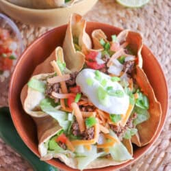 Taco salad topped with sour cream & green onions in a tortilla bowl in an orange bowl on a wooden table.