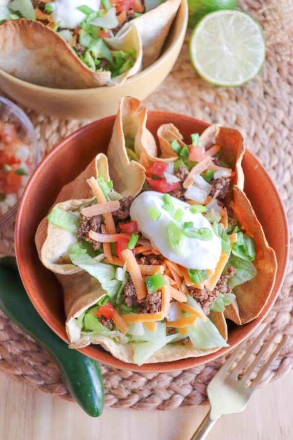 Taco salad topped with sour cream & green onions in a tortilla bowl in an orange bowl on a wooden table.
