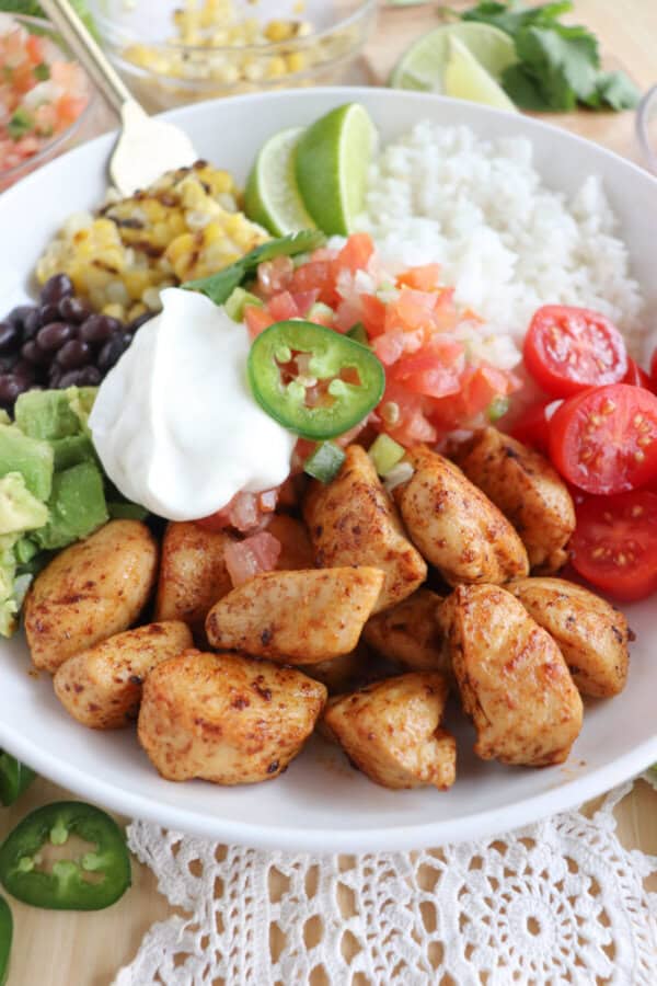 Cooked chicken bites with seasoning in a large white bowl with rice, diced avocado, black beans, corn kernels, cherry tomatoes, pico and sour cream topped with a slice of jalapeno and lime.